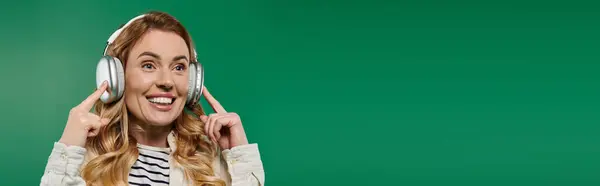 A cheerful woman in casual attire listens to music, smiling brightly against a vivid green backdrop. — Stock Photo