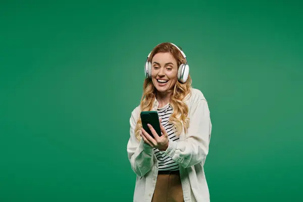 A curly-haired woman dressed casually smiles brightly as she engages with her phone, lost in music. — Stock Photo