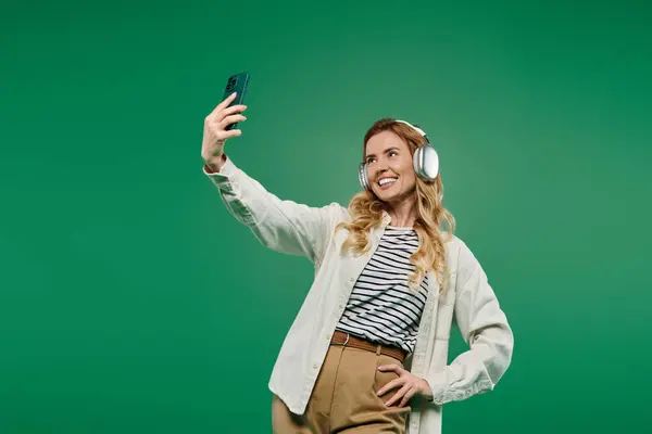 A joyful woman in casual wear takes a selfie with headphones, set against a bright green background. — Stock Photo