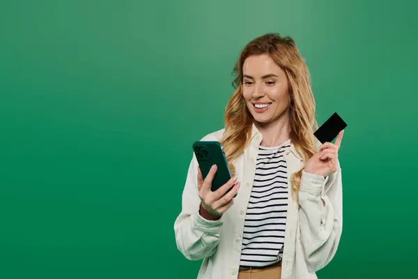 A smiling woman with curly hair holds a smartphone and a card in a bright green environment, radiating joy. — Stock Photo