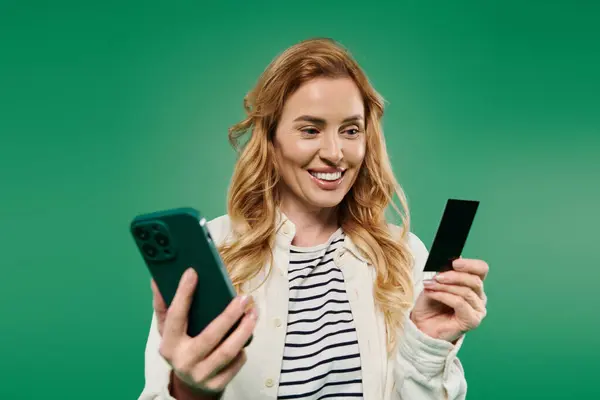 A cheerful woman with curly hair holds her phone and a card, exuding excitement and engagement in a vibrant setting. — Stock Photo