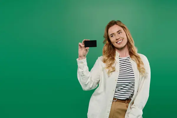 A woman with curly hair smiles brightly while holding a card, dressed casually against a green backdrop. — Stock Photo