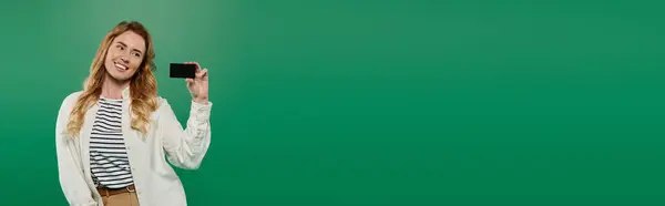 A cheerful woman in casual attire smiles brightly while holding a smartphone on a vivid green background. — Stock Photo