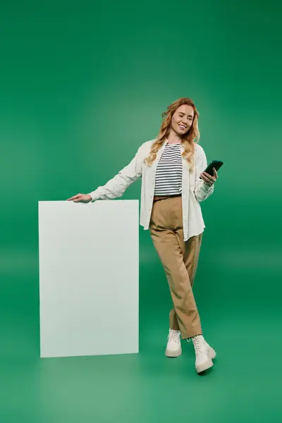A smiling woman stands casually with a smartphone in hand against a vibrant green background, conveying joy. — Stock Photo