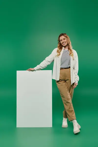 Uma mulher alegre em traje casual fica ao lado de uma tela em branco, irradiando calor e criatividade. — Fotografia de Stock