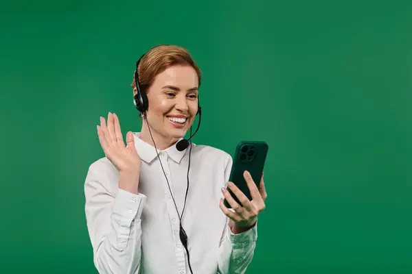 A woman in formal wear smiles and waves while interacting on her phone against a bright green background. — Stock Photo