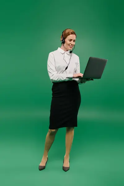 A confident woman in a white shirt and skirt uses a laptop to engage with clients against a green background. — Stock Photo