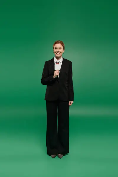 A confident TV presenter engages the audience in black formal wear, set against a lively green backdrop. — Stock Photo