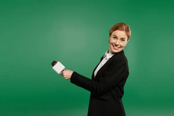 A confident TV presenter in a stylish black suit interacts warmly with the audience against a lively green background. — Stock Photo