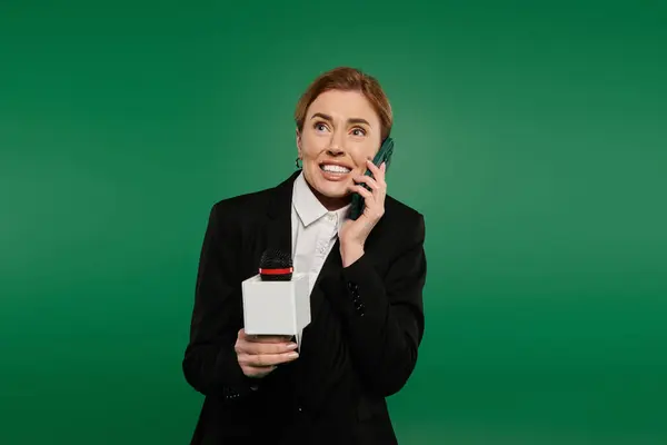 A TV presenter in formal black chats enthusiastically on the phone against a green backdrop. — Stock Photo