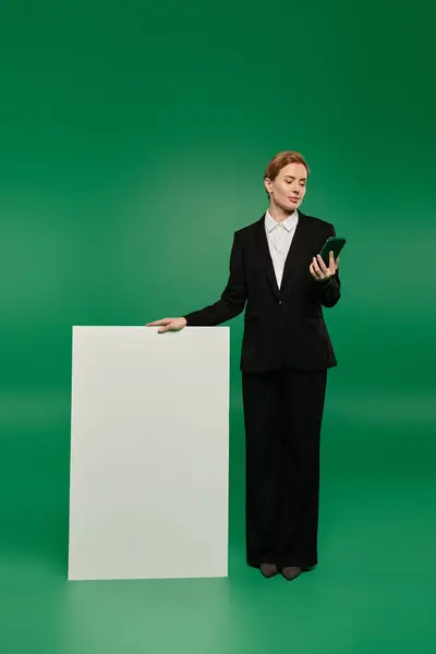 A stylish TV presenter in black holds a smartphone, poised to engage viewers on a lively green set. — Stock Photo