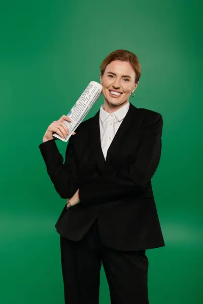 A confident TV presenter in black formal attire smiles while holding a newspaper against a vivid green backdrop. — Stock Photo