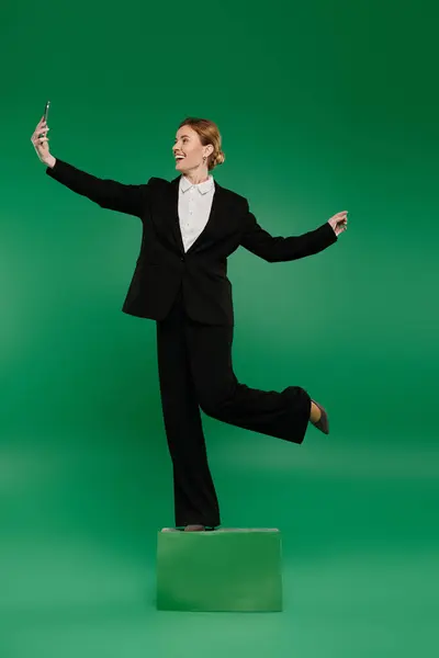 A stylish TV presenter in black formal attire interacts playfully while standing on a pedestal, surrounded by green. — Stock Photo
