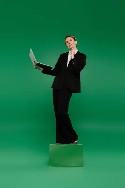 A stylish TV presenter stands confidently on a platform, pondering while holding a laptop amidst a green setting. — Stock Photo