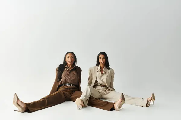 Two young women showcase fashionable attire while seated on a minimalistic background. — Stock Photo