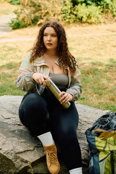 Una hermosa mujer de talla grande se sienta en una roca, tomando un descanso en un bosque soleado rodeado de vegetación. - foto de stock