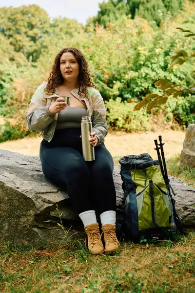 Une femme de taille plus savoure du thé chaud tout en se reposant sur un rocher au milieu d'une verdure luxuriante. — Photo de stock