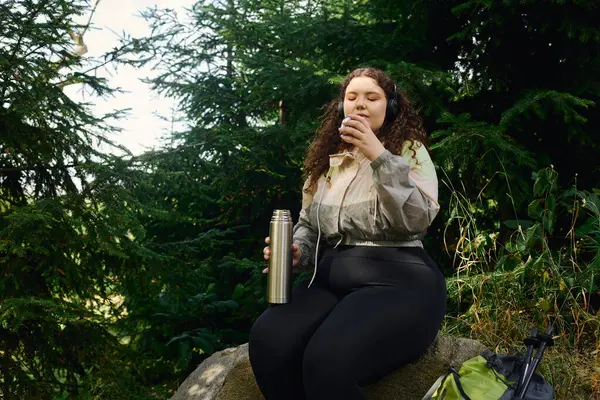 A vibrant plus size woman savors a warm drink in a tranquil green forest surrounded by trees. — Stock Photo