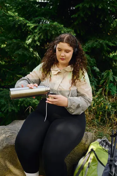 A beautiful plus size woman sits on a rock, savoring a hot drink surrounded by lush greenery. — Stock Photo