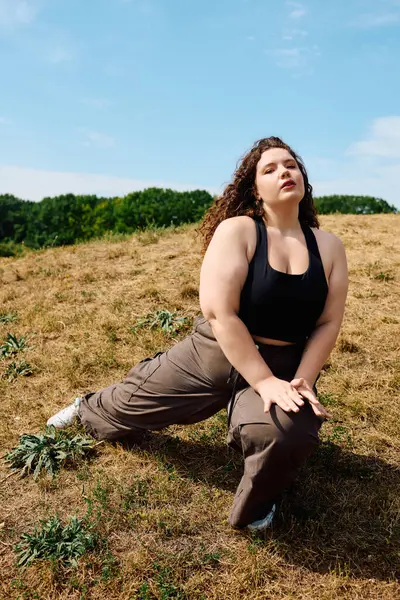 A beautiful plus size woman gracefully stretches on a grassy hillside under a clear blue sky. — Stock Photo