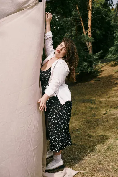 A confident plus size woman playfully interacts with a fabric backdrop in a serene setting. — Stock Photo