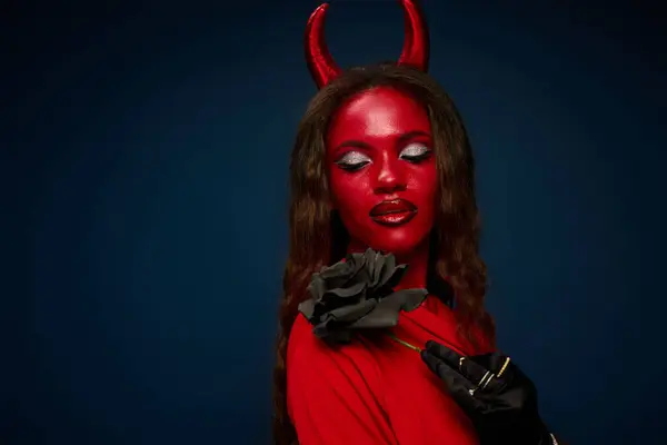 The captivating woman in red devil attire holds a dark rose, embodying Halloween spirit. — Stockfoto