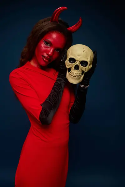A beautiful woman dressed as a devil holds a skull, capturing the Halloween spirit beautifully. — Stock Photo