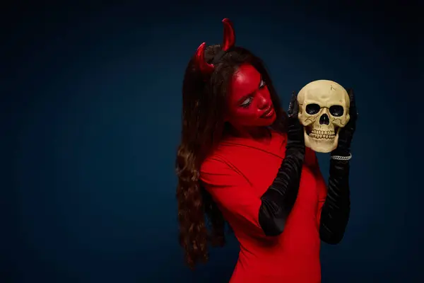 A stunning woman in a devil outfit passionately examines a skull, embracing Halloween spirit. — Stock Photo