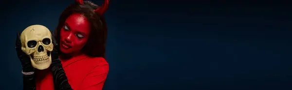 A beautiful woman dressed as a devil admires a skull in a dark, moody setting for Halloween. — Stock Photo