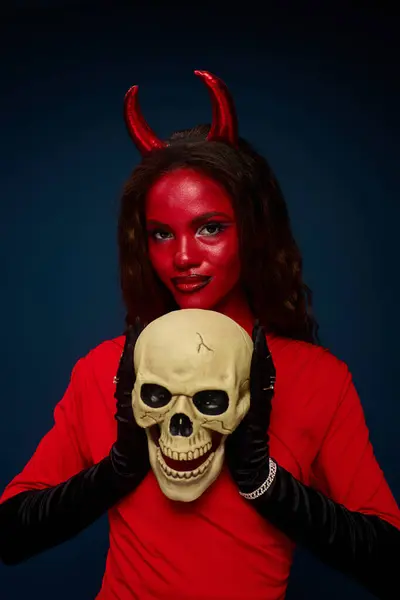 A captivating woman dressed as a devil holds a skull, showcasing Halloween spirit and creativity. — Stock Photo