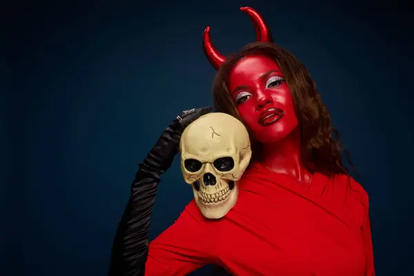 A captivating woman dressed as a devil holds a skull, embodying Halloween spirit and allure. — Stock Photo