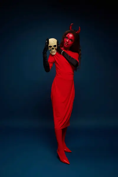 Uma mulher deslumbrante vestida como um diabo graciosamente posa com um crânio, abraçando o espírito de Halloween. — Fotografia de Stock