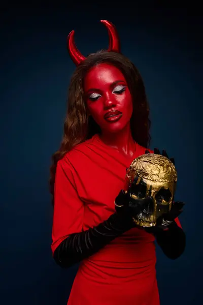 In a vivid red outfit, a stunning woman embraces Halloween spirit while holding a golden skull. — Stock Photo