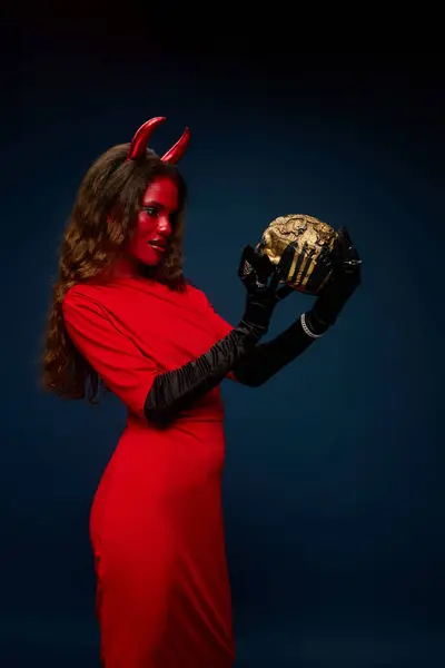 A stunning woman dressed as a devil admires a gleaming golden skull in an enchanting atmosphere. — Stock Photo