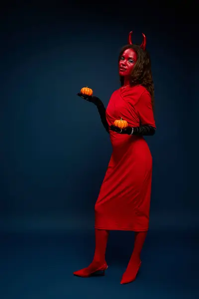 Uma mulher deslumbrante vestida com trajes vermelhos mostra seu espírito de Halloween com abóboras na mão. — Fotografia de Stock