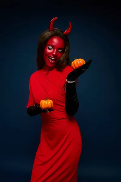 A bewitching woman in a devil outfit holds two pumpkins, embodying Halloween spirit. — Stock Photo