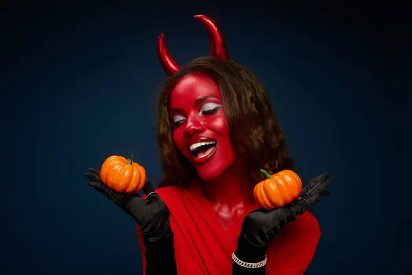 A beautiful woman dressed as a devil joyfully showcases two pumpkins while celebrating Halloween. — Stock Photo
