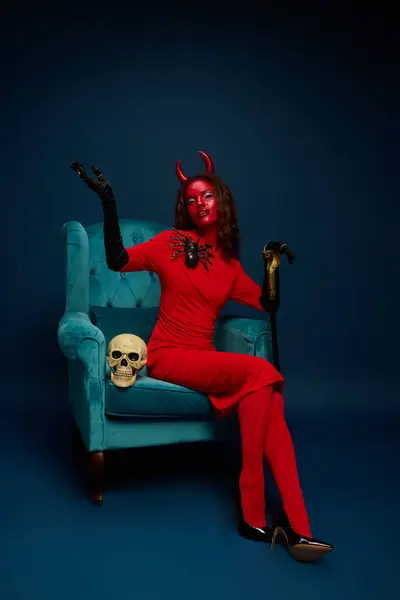 A stunning woman in a devil outfit confidently sits in a chic turquoise chair for Halloween. — Stock Photo