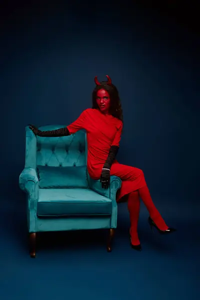 The enchanting woman, dressed in a vivid red costume, strikes a pose on a teal armchair. — Stock Photo