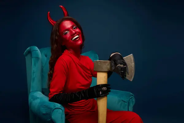 La femme frappante dans un costume du diable sourit lumineux, tenant une hache tout en étant assis dans une chaise élégante. — Photo de stock