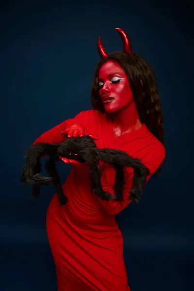 A stunning woman dressed in red with horns plays with a spider prop at a Halloween event. — Stock Photo