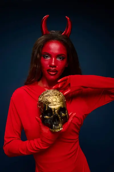 The captivating woman in a red devil costume poses confidently, clutching a golden skull. — Stock Photo