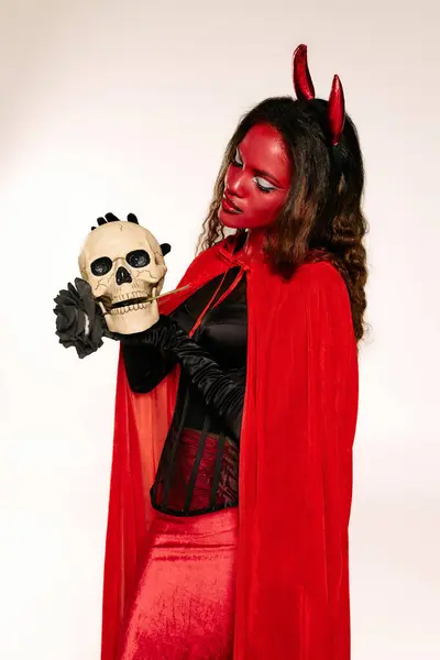 The alluring woman in a red devil costume holds a skull, radiating Halloween spirit. — Stock Photo