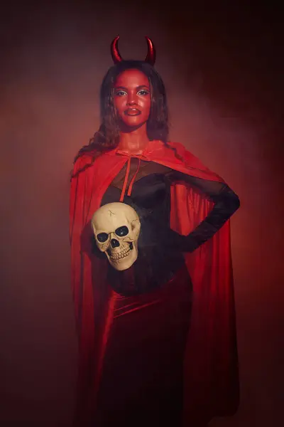 A striking woman dressed as a devil poses confidently, showcasing her Halloween spirit with a skull. — Stock Photo