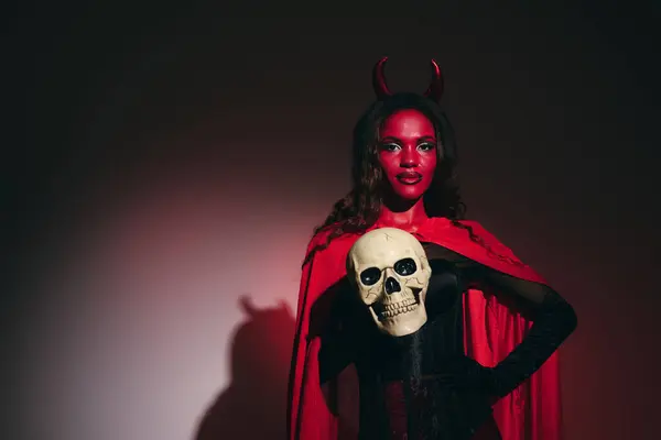 Dressed as a devil, she confidently holds a skull, celebrating the spirit of Halloween. — Stock Photo