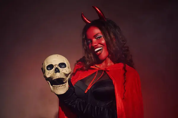 A striking woman dressed as a devil expresses joy while holding a skull, celebrating Halloween. — Stock Photo