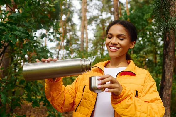 Una joven vierte una bebida caliente de su termo mientras camina en el vibrante bosque otoñal, abrazando la naturaleza. - foto de stock