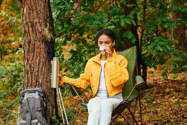 In a colorful autumn forest, a young woman relaxes with a warm drink, surrounded by vibrant foliage. — Stock Photo