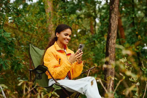 A young woman dressed in vibrant autumn colors smiles as she checks her smartphone in the forest. — Stock Photo