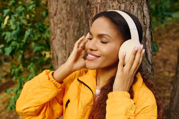 A young African American woman smiles while hiking through an autumn forest, listening to music with joy. — Stock Photo